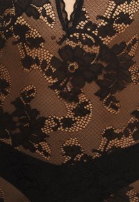 Svart blonde bodysuit med blomster mønster, gjennomsiktig tekstur og dyp utringning. Myk stoff med blomster detaljer og en kontrasterende bunntrim.