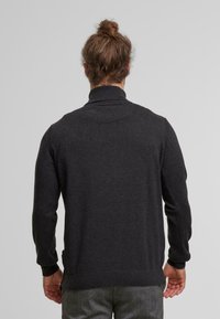 Mann mit braunem, zusammengebundenem Haar, der einen dunkelgrauen, langärmeligen Pullover und graue Hosen trägt und mit dem Rücken zu einem schlichten, hellen Hintergrund steht.