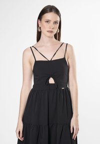 El vestido negro sin mangas presenta un corsé ajustado con un detalle de ojal en la parte delantera y una falda de volantes en forma de capas, confeccionado en un tejido suave.