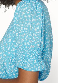 Blusa floreale blu con maniche corte a sbuffo, raccolte ai polsini. Realizzata in tessuto leggero, presenta motivi floreali bianchi su tutta la superficie.