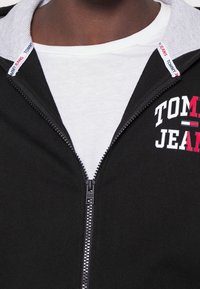 Felpa nera con zip e fodera grigia nel cappuccio, con testo "TOMMY JEANS" bianco e rosso. Sotto c'è una maglietta bianca. Tessuto con texture liscia.