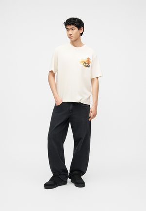 Bărbat tânăr în picioare, purtând un tricou alb cu mâneci scurte, având un mic grafic colorat, și blugi negri largi cu adidași negri.