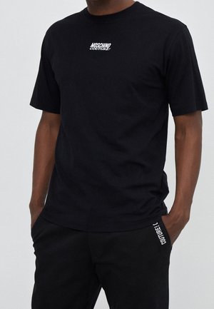 Hombre con camiseta negra Moschino Couture y pantalones negros a juego, con las manos en los bolsillos, sobre un fondo claro y liso.