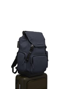 Un sac à dos bleu marine avec plusieurs poches zippées, des bretelles réglables et un système de fermeture à rabat, reposant sur une valise vert foncé. Matériau texturé.