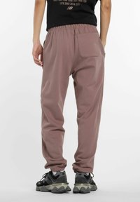 Man in mauve joggerbroeken met elastische manchetten en een zwart T-shirt, staand met één hand in de broekzak, zichtbaar van achteren.