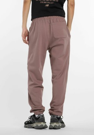 Man in mauve joggerbroeken met elastische manchetten en een zwart T-shirt, staand met één hand in de broekzak, zichtbaar van achteren.