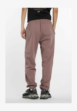 Uomo che indossa pantaloni jogger color malva con polsini elasticizzati e una t-shirt nera, in piedi con una mano nella tasca dei pantaloni, visibile da dietro.