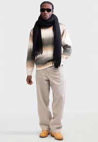 Pull en tricot avec des rayures dégradées beige, vertes et brunes, associé à un pantalon beige clair et une écharpe noire à franges. Bottines marron à enfiler.