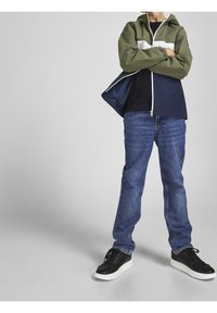 Chaqueta cortavientos en bloque de color verde y blanco con capucha, combinada con jeans de mezclilla azul y zapatillas negras con suelas blancas.