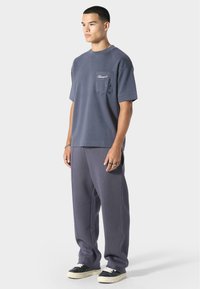 T-shirt oversize gris avec une poche sur la poitrine, associé à un pantalon de jogging gris assorti avec des coutures latérales et des chaussures noires avec des semelles blanches.