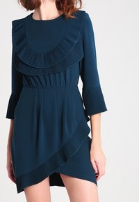 Teal klänning med en veckad volangkrage, trekvartsärmar med veck och en asymmetrisk fåll, tillverkad av mjukt tyg.