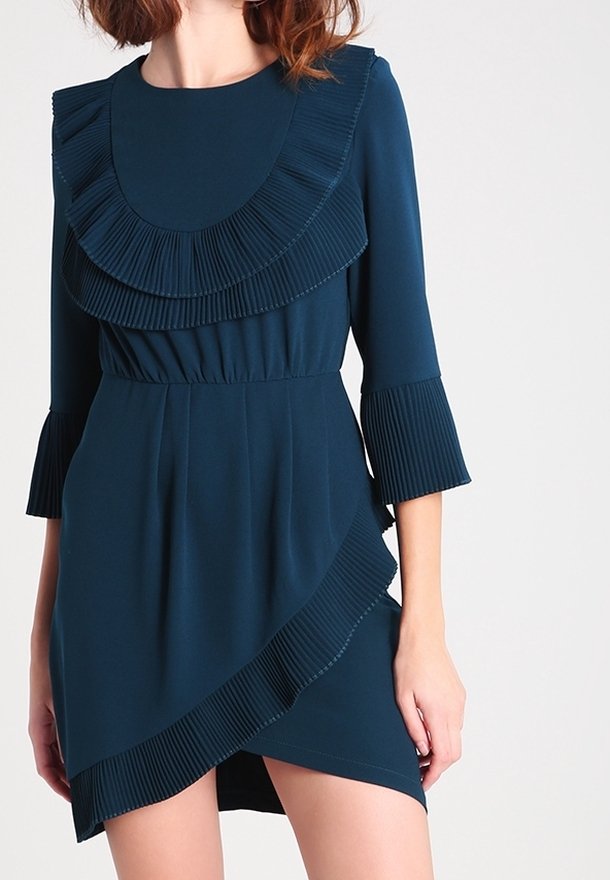 Teal klänning med en veckad volangkrage, trekvartsärmar med veck och en asymmetrisk fåll, tillverkad av mjukt tyg.