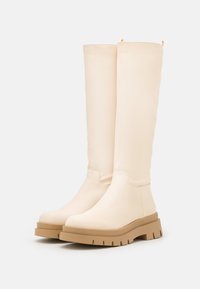 Botas altas até ao joelho, de cor creme, feitas de material liso, com sola espessa bege e padrão texturizado para melhor aderência. Apresenta uma alça lateral para facilitar o calçar.