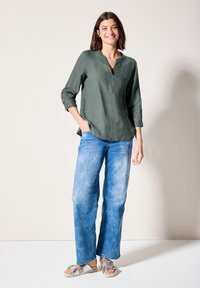 Camicia di lino verde oliva con scollo a V e maniche a tre quarti, abbinata a jeans a gamba larga blu chiaro e sandali grigi con un dettaglio a fiocco.