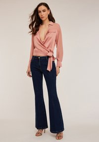 Blusa de satén rosa con mangas largas y un lazo en la cintura, combinada con jeans de campana azul oscuro y tacones atados.