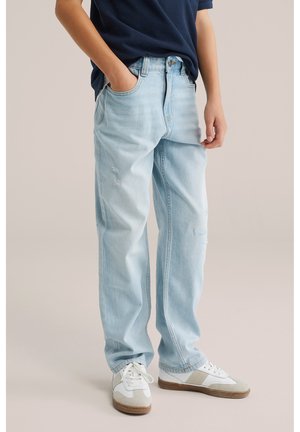 Kind draagt lichtblauwe gescheurde jeans, marineblauw shirt en witte sneakers met beige accenten, staand met één hand in de zak.