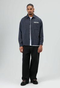 Geruit marineblauw overhemdjack met knoopsluiting en zak, gedragen over een wit overhemd, gecombineerd met zwarte wijde broekspijpen en zwarte schoenen.