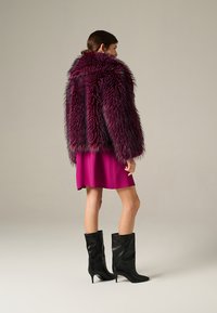 Giacca in pelliccia sintetica di un profondo viola con una texture lunga e shaggy, abbinata a un vestito rosa e stivali neri con tacco, vista di spalle.