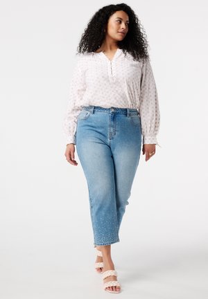Blouse blanche avec un motif subtil, manches longues et fermeture à boutons, associée à un jean bleu clair avec des poignets embellis et des sandales à glisser.