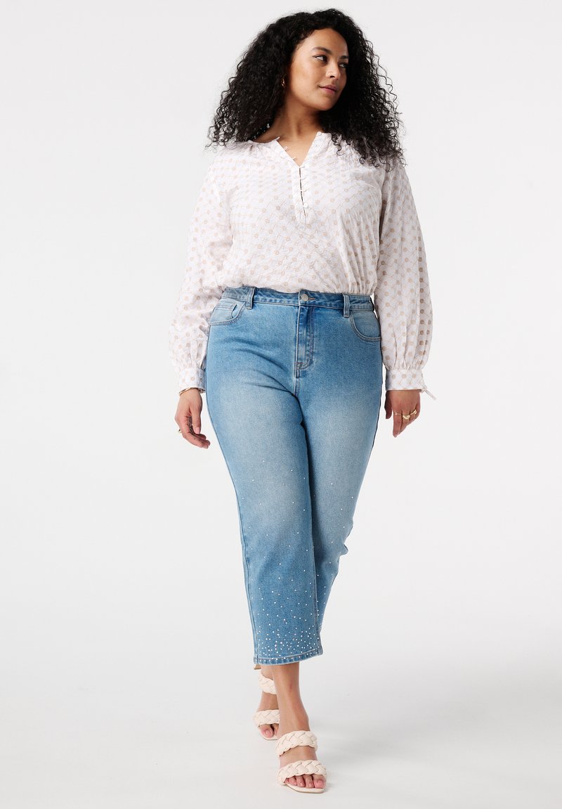 Blouse blanche avec un motif subtil, manches longues et fermeture à boutons, associée à un jean bleu clair avec des poignets embellis et des sandales à glisser.