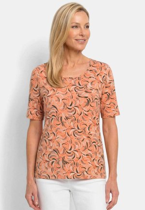 Frau mit glattem blonden Haar, die ein kurzärmeliges Pfirsichfarbenes Shirt mit abstrakten schwarzen und pinken Wirbelmustern trägt, kombiniert mit weißen Hosen.