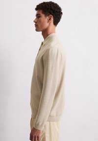 Beiger Strick-Polo-Pullover mit langen Ärmeln, umgeschlagenem Kragen, Drei-Knopf-Leiste und gerippten Bündchen und Saum. Glatte Textur, taillierte Form.