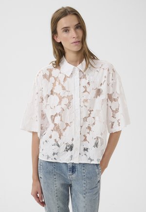 Młoda kobieta ubrana w białą, kwiatową, koronkową koszulę na krótki rękaw zapinaną na guziki, na ciemny top, zestawioną z jasnoniebieskimi, wysokim stanem jeansami.