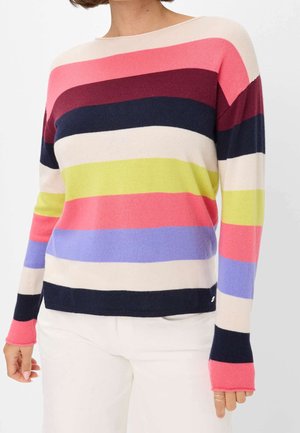Femme portant un pull à manches longues avec de larges rayures horizontales en rose, bordeaux, marine, crème, lime et lavande, associé à un pantalon blanc.