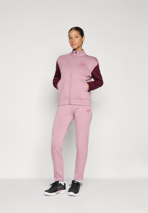 Nő rózsaszín melegítőben, bordó ujjakkal és fekete sportcipőben, zsebeiben tartva a kezét, egyszerű fehér háttér előtt áll.