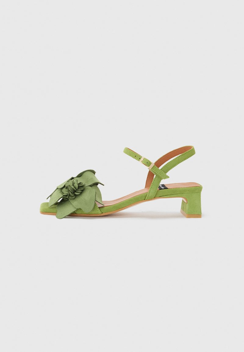 Sandal in suede verde con cinturino alla caviglia regolabile, punta quadrata e dettaglio floreale a strati sulla parte anteriore. Tacco basso a blocco.