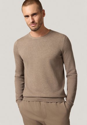 Beżowy sweter z długim rękawem, wykonany z dzianiny, z okrągłym dekoltem, prążkowanymi mankietami i dołem. Miękka struktura z delikatnym wzorem, w zestawieniu z beżowymi spodniami.