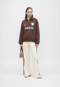 Rjav oversized pulover s turkiznim logotipom Adidas, v kombinaciji s smetanastimi širokimi hlačami z maroon stranskimi črtami. Majhna črna torbica.