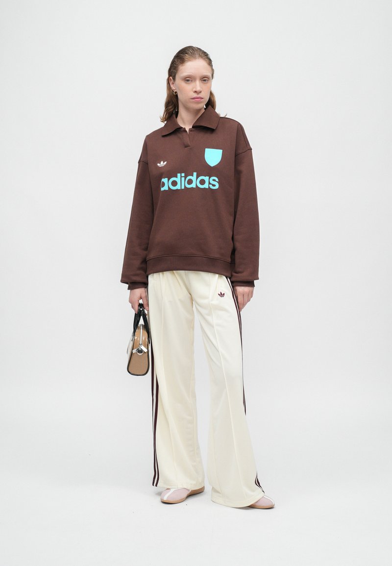 Sweatshirt surdimensionné marron avec logo Adidas turquoise, associé à un pantalon ample crème avec des rayures latérales bordeaux. Petit sac à main noir.