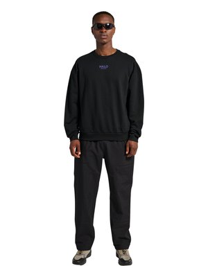 Man met een zwarte sweater met de tekst "HALO", zwarte broek, zonnebril en beige sneakers, staand tegen een witte achtergrond.