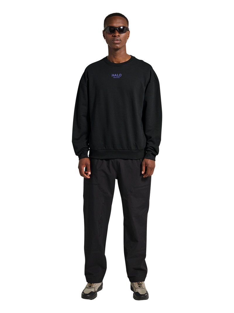 Mężczyzna w czarnej bluzie z napisem „HALO”, czarnych spodniach, okularach przeciwsłonecznych i beżowych butach sportowych stoi na białym tle.