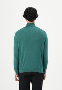 Person mit kurzen lockigen dunklen Haaren, die einen türkisfarbenen Langarmrollkragenpullover und schwarze Hosen trägt, mit dem Rücken zu einer schlichten Hintergrund.