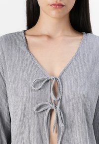 Light gray textured blouse with a deep V-neck, featuring a tie closure at the neckline and loose, flowing sleeves.
