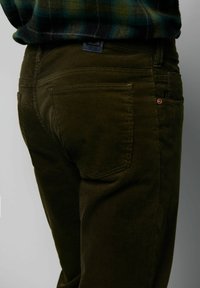 Spodnie sztruksowe w ciemnooliwkowym kolorze, charakteryzujące się subtelną prążkowaną teksturą. Ważne detale to tylne kieszenie oraz mała metka na pasku talii.