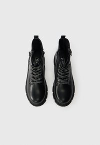 Par črne usnjene gležnjarje z debelimi podplati, vzorčastimi črnimi in belimi vezalkami ter stranskimi zadrgami na beli podlagi.