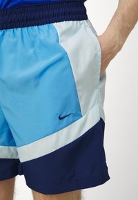 Blå og lyseblå svømmebukser med elastisk linning, sidelommer og en liten marineblå logo på venstre lår. Teksturert stoffpaneler.