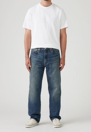 Mand iført en ensfarvet hvid t-shirt med korte ærmer, blå afslappede denimjeans og off-white snørebåndssneakers, stående mod en neutral baggrund.