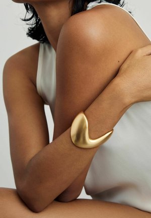 Inimene, kes kannab valget varrukateta topi ja kelle kõverdatud käel on sile kuldne käevõru, mis toetub rinnale.