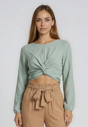 Frau mit langen hellbraunen Haaren, die ein hellgrünes gedrehtes Crop-Top und beige hoch taillierte Hosen mit gebundenem Gürtel trägt und vor einem schlichten Hintergrund posiert.