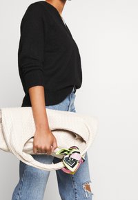 Schwarzer Pullover mit V-Ausschnitt, kombiniert mit hellblauen, verwaschenen Jeans. Beigefarbene, gewebte Tasche mit einem bunten Schal, der am Griff gebunden ist.