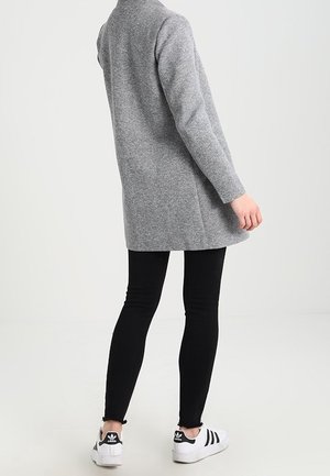 Grauer langer Mantel mit einer weichen Textur, getragen über schwarzen Skinny Jeans und weißen Sneakers mit schwarzen Streifen. Einfaches, schlankes Design.