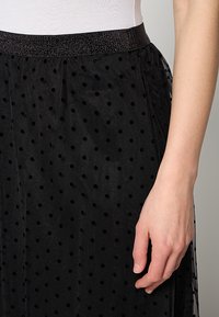 Falda negra hecha de tejido transparente con lunares texturizados. Cuenta con una cinturilla elástica ancha y brillante, y un diseño fluido.