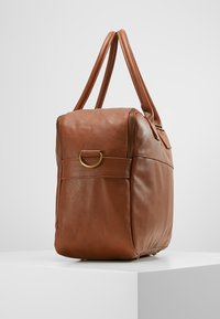 Borsa in pelle marrone con una superficie liscia, doppi manici superiori e un accento in metallo dorato. Presenta una chiusura a zip e una forma arrotondata.