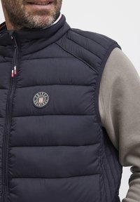Mornarsko pufasta prsluk s horizontalnim šavovima, zatvaranjem na zip i okruglom logotipom na prsima. Nosi se preko svijetlosivog dugog rukava.