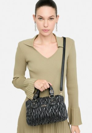 Borsa a tracolla nera trapuntata con doppio manico, dettagli in oro e tracolla staccabile, tenuta da un modello che indossa un vestito oliva a coste.