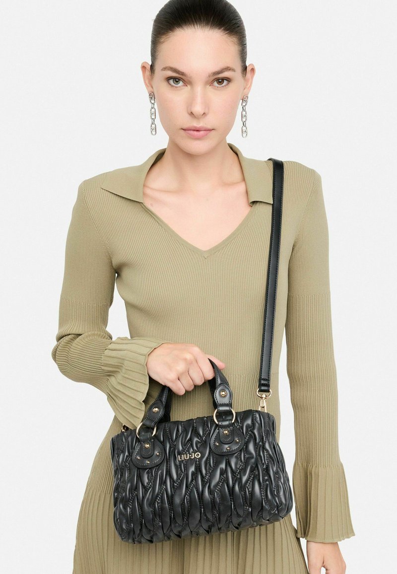 Borsa a tracolla nera trapuntata con doppio manico, dettagli in oro e tracolla staccabile, tenuta da un modello che indossa un vestito oliva a coste.